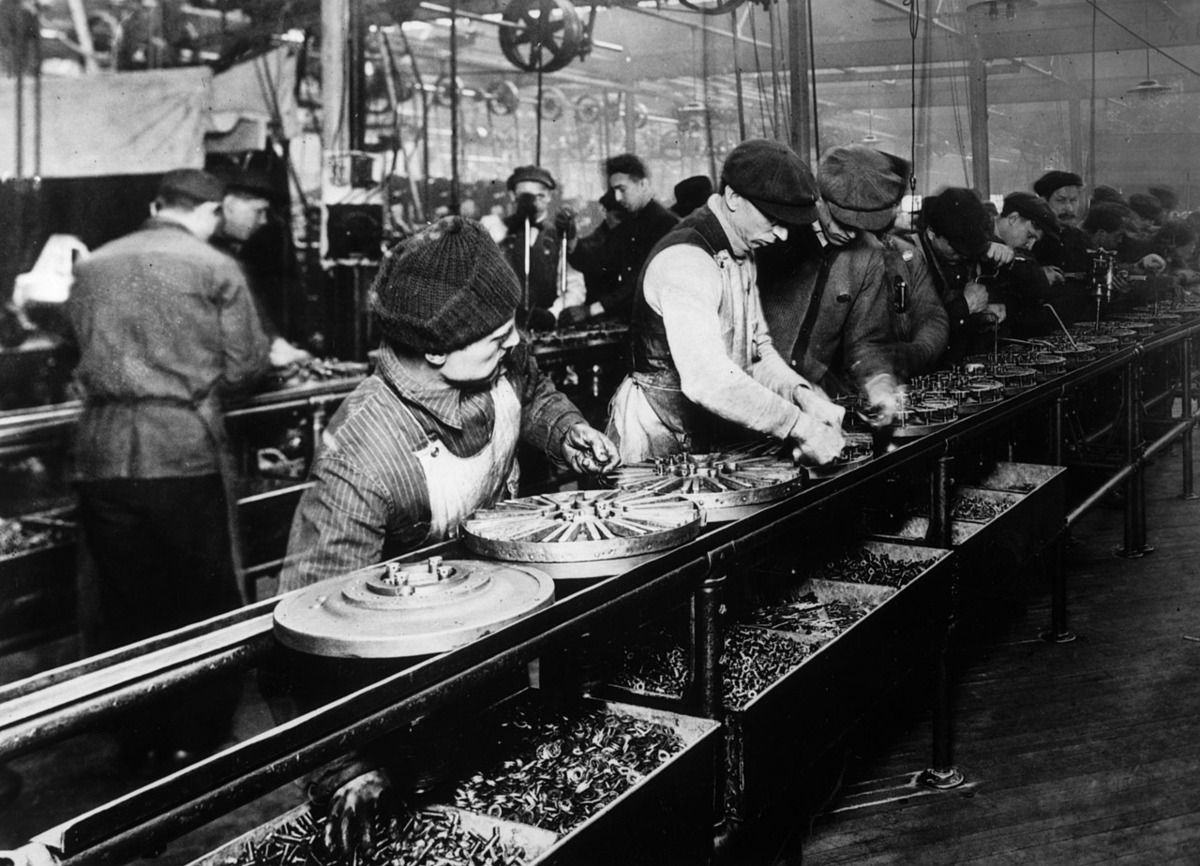 Early 1900s factory assembly line. Workers in caps assemble mechanical parts on conveyor belts. Industrial setting with machinery visible